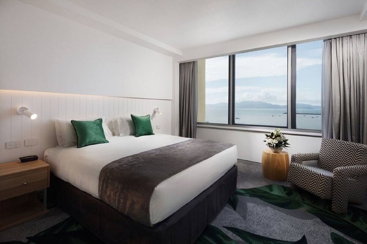 A hotel room with a large bed, a nightstand, a chair, a window with a view of the sea, and a small table with a plant.