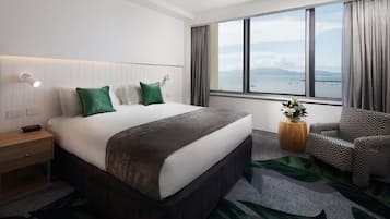 A hotel room with a large bed, a nightstand, a chair, a window with a view of the sea, and a small table with a plant.