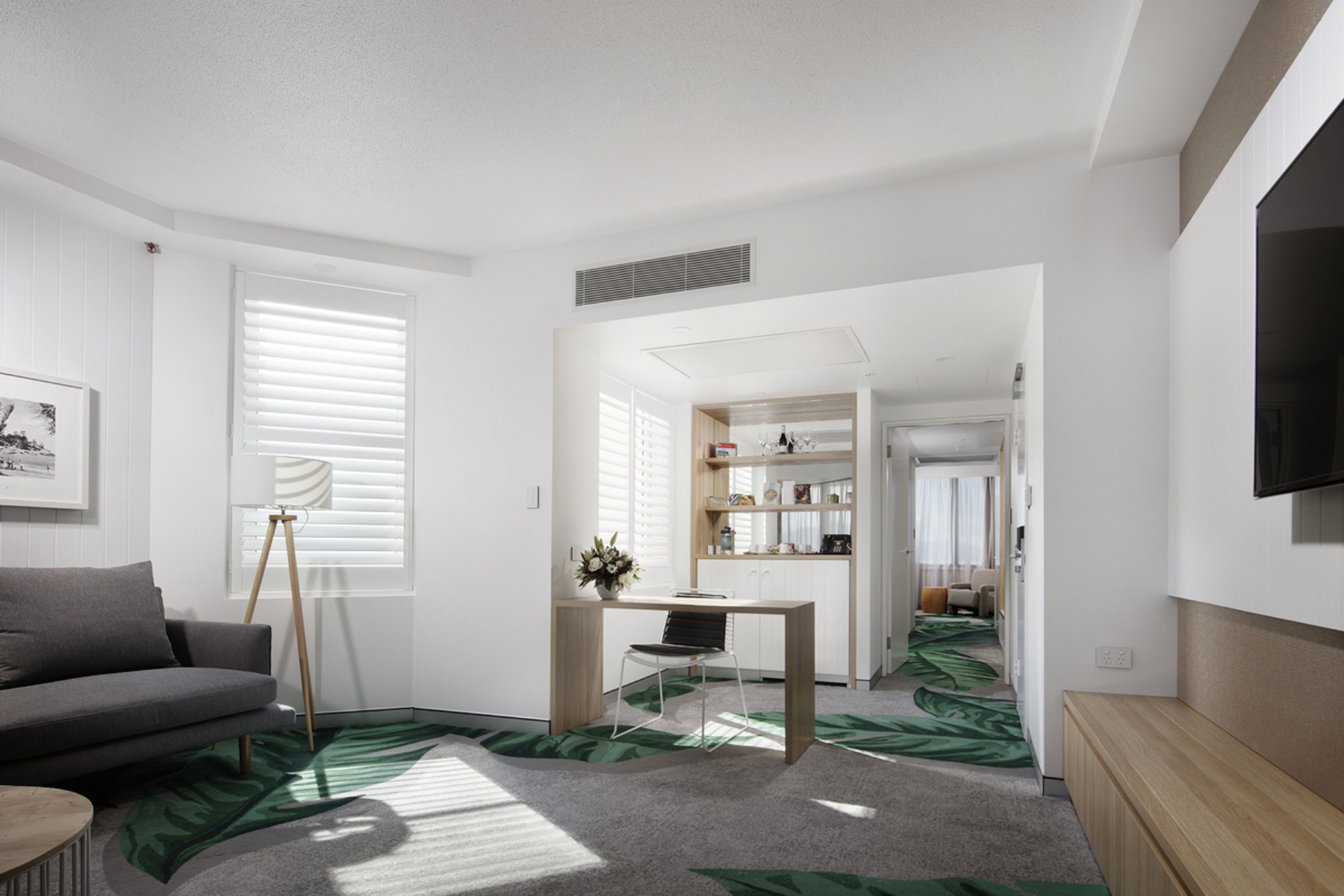 A modern living room with a grey sofa, a wooden coffee table, and a dining area with a wooden table and chairs. There is a TV mounted on the wall and a window with blinds.