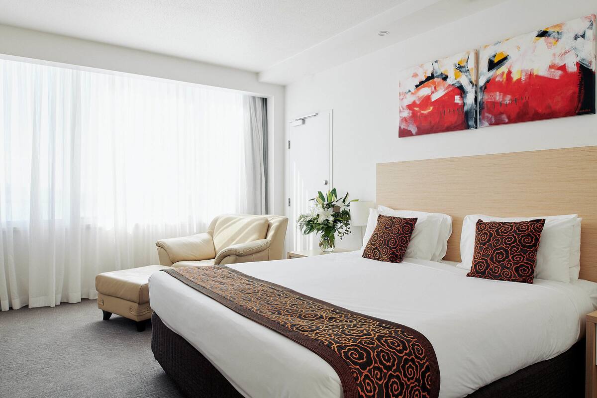A hotel room with a large bed, a chair, a bedside table, and a painting on the wall.