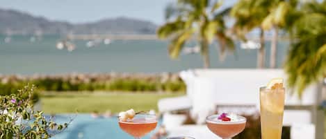A terrace with a pool view, three drinks, and a white railing.