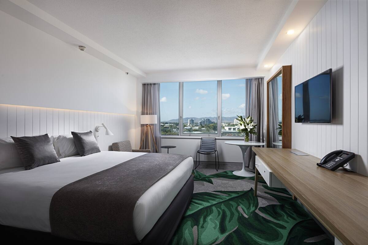 A modern hotel room with a large bed, a desk, a chair, a flat-screen TV, and a view of the city.