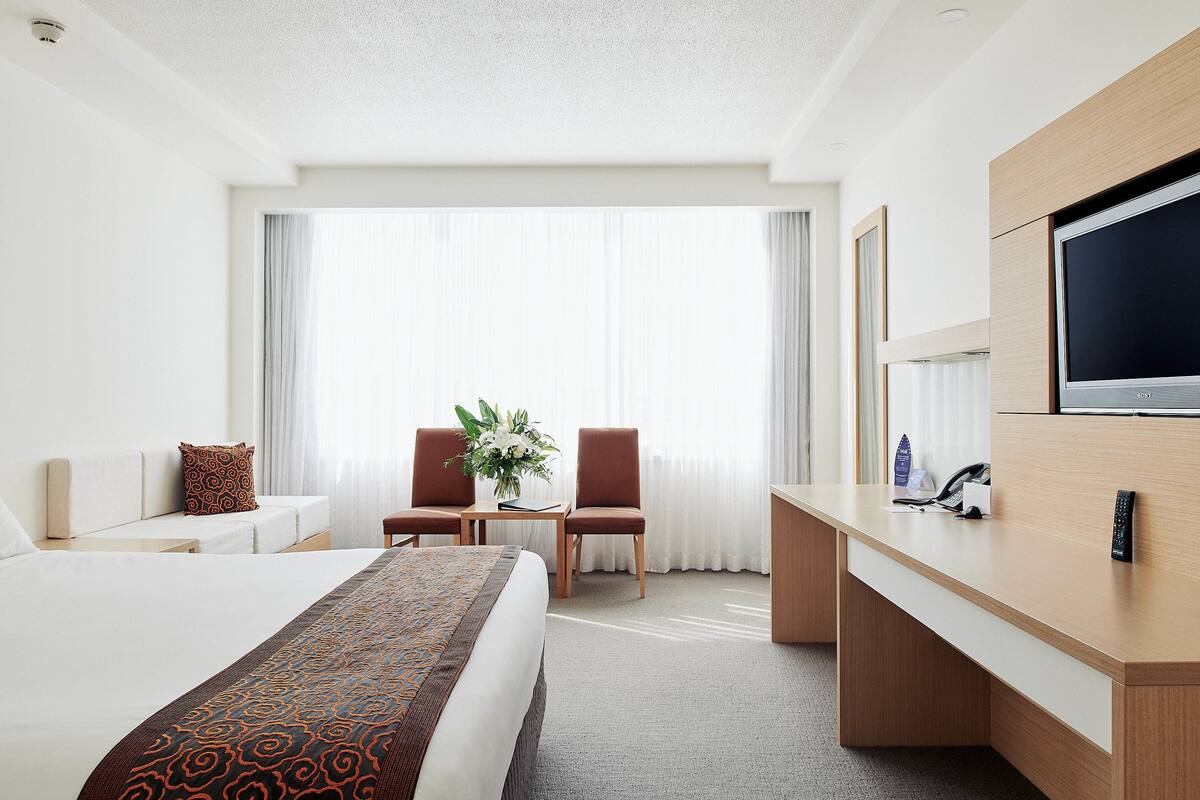 A hotel room with a large bed, a desk with a flat-screen TV, two chairs, a small table with a vase of flowers, and a large window with curtains.