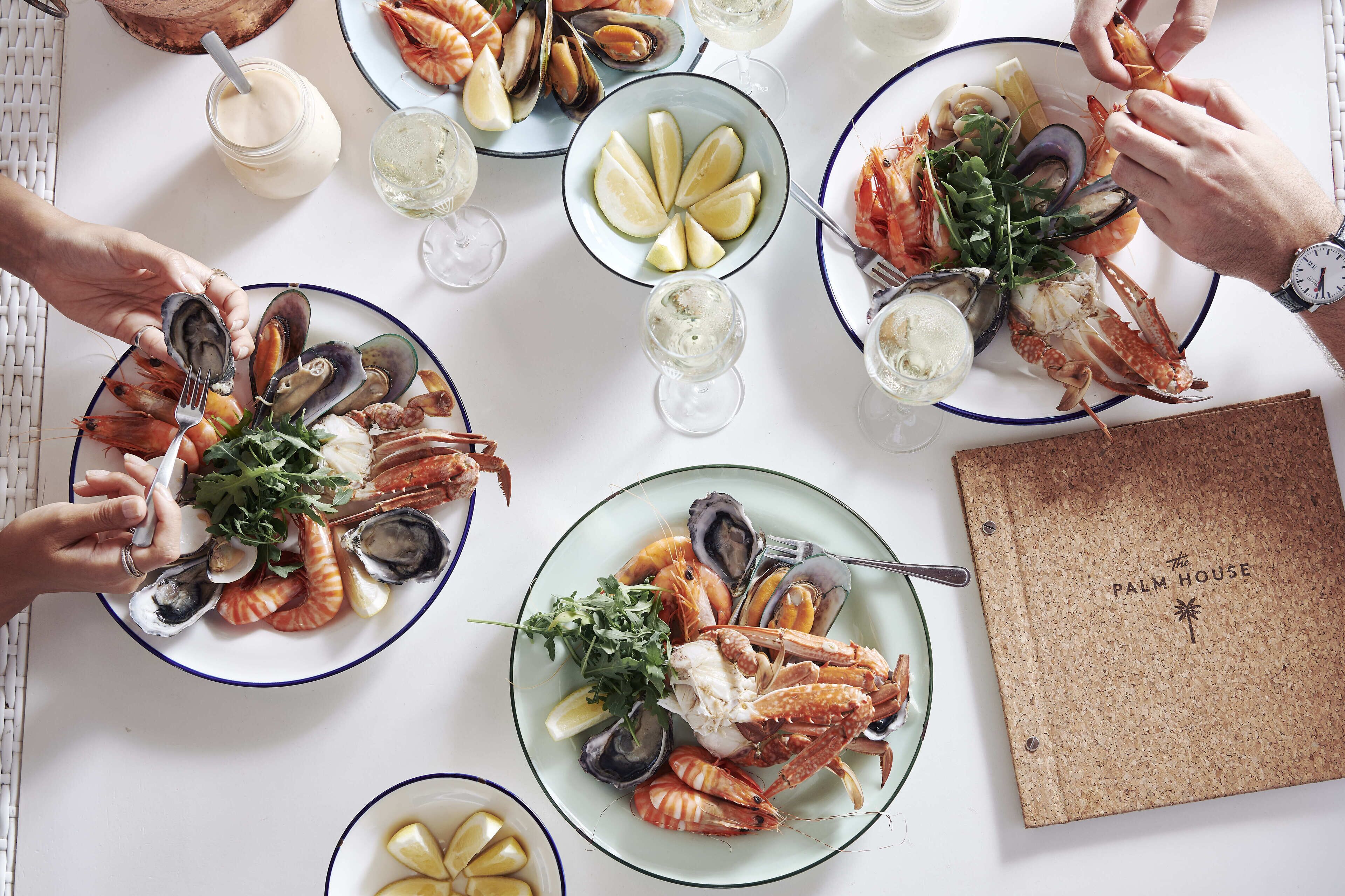 Une table dressée avec des plats de fruits de mer, des verres de vin blanc et un tableau d’affichage en liège sur lequel on peut lire : « The Palm House ».