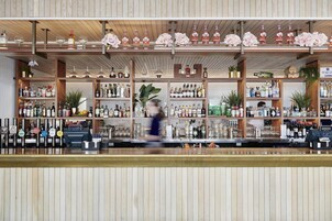 Zona de bar con estanterías de madera llenas de botellas y vasos, barra y una persona en movimiento.