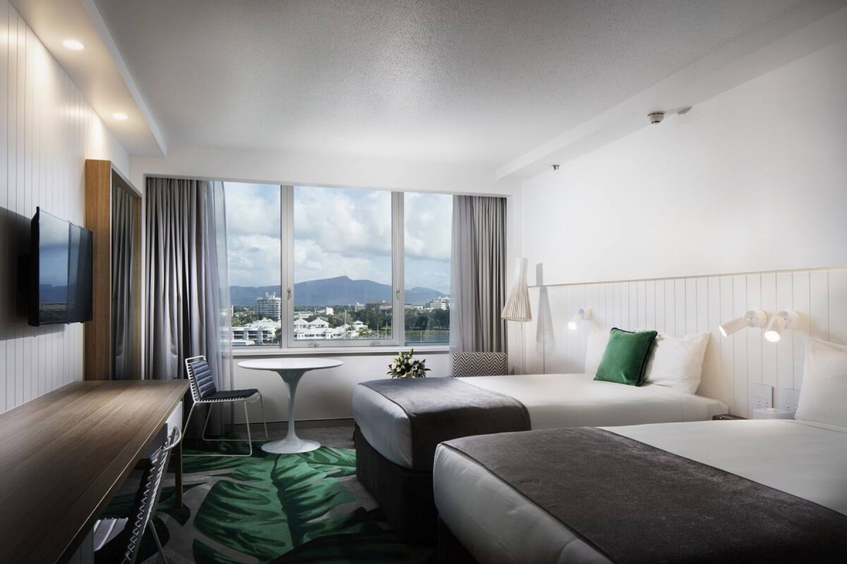 A hotel room with two beds, a desk, a TV, and a view of the city and mountains.