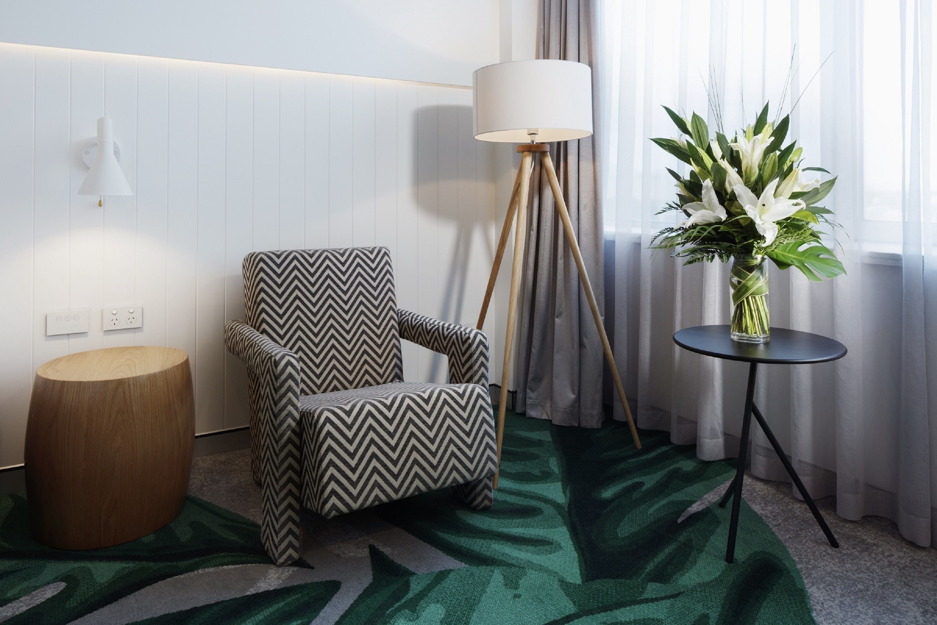 Una habitación moderna con un sillón de diseño en zigzag, una mesita redonda de madera, una lámpara de pie y un jarrón con flores blancas.