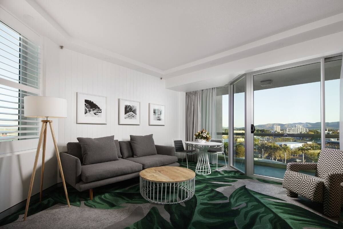 A modern living room with a grey sofa, a round wooden coffee table, a floor lamp, and a view of the city through large windows.