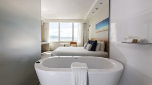 A modern hotel room with a large bathtub, a bed with pillows, a desk, and a view of the sea.