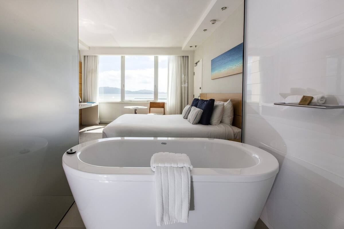 A modern hotel room with a large bathtub, a bed with pillows, a desk, and a view of the sea.
