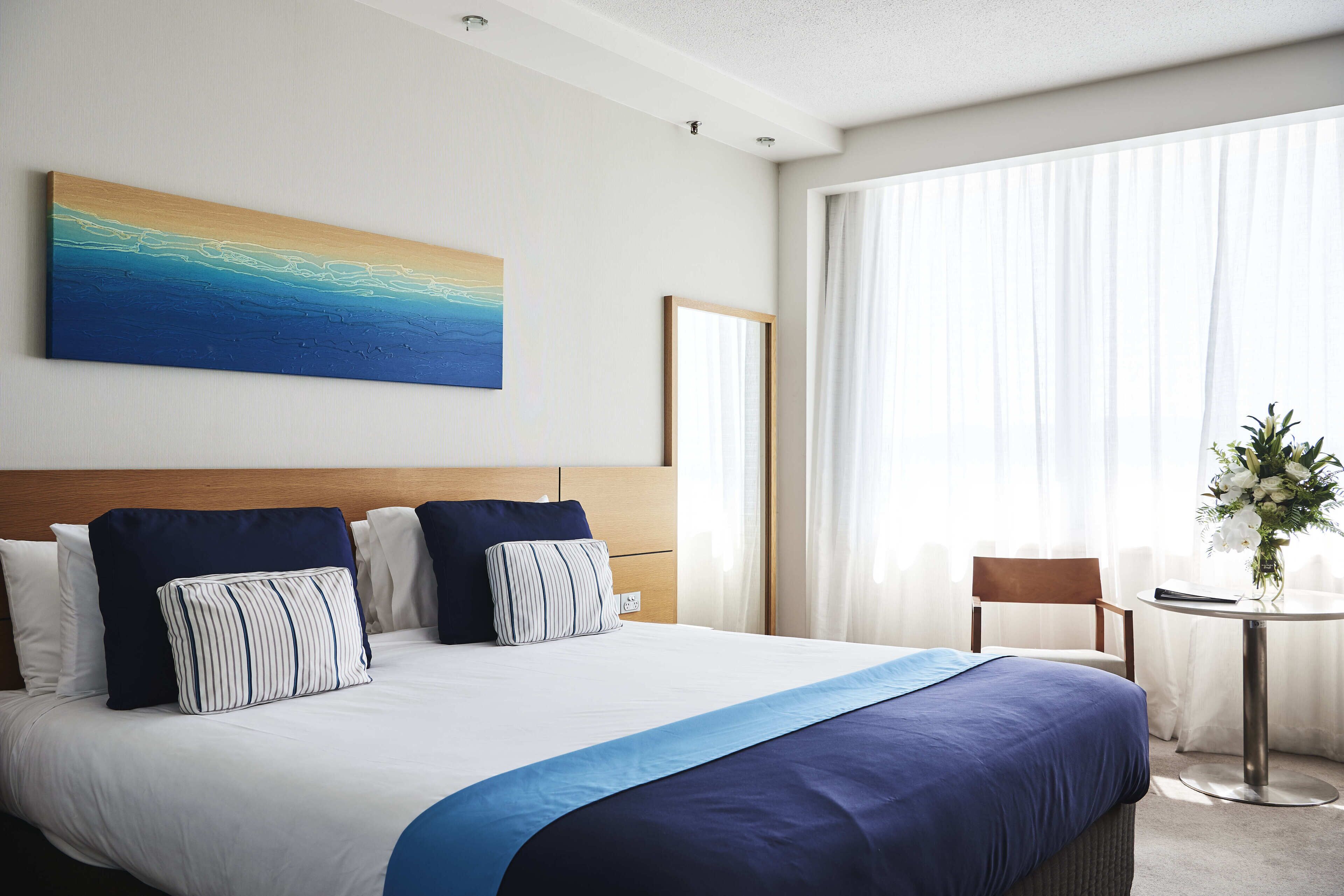 A neatly made bed with a blue and white bedspread, a wooden headboard, and a large abstract painting above it.
