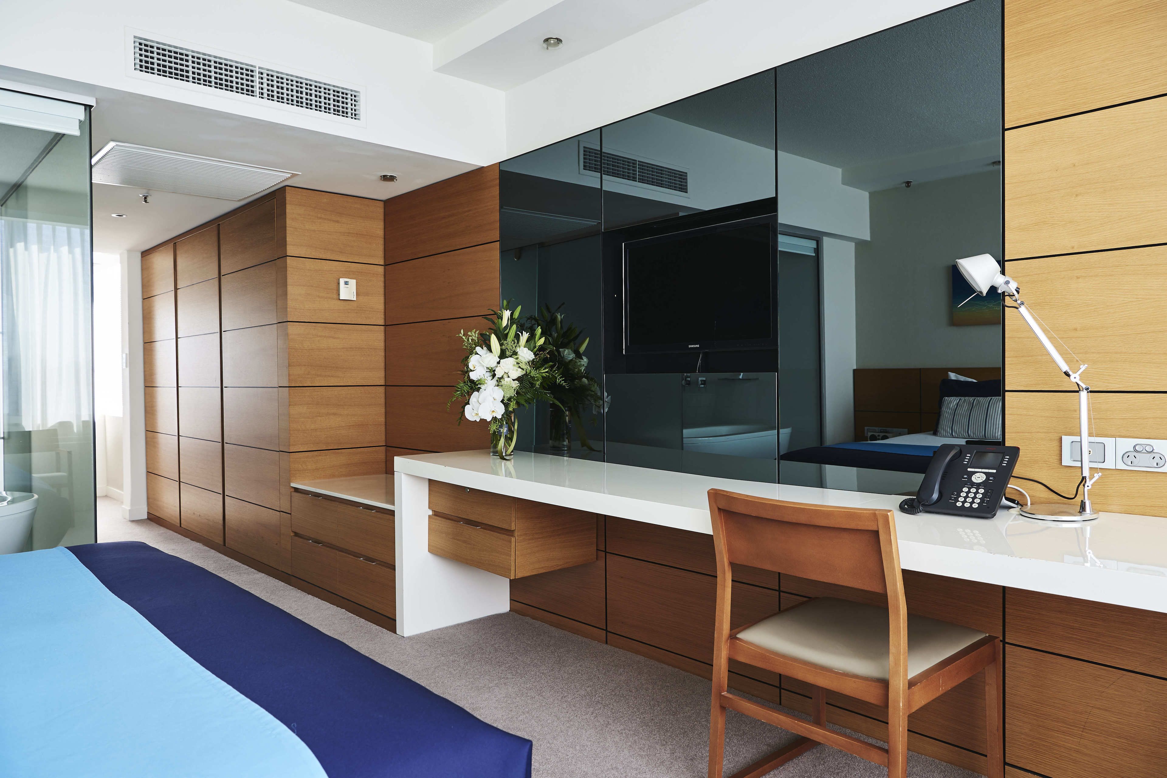 Une chambre d’hôtel moderne avec un grand lit, un bureau en bois, une chaise, une lampe de bureau, un téléphone et un vase avec des fleurs.