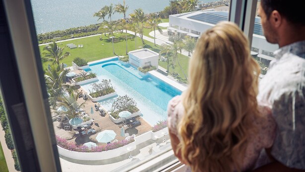 A view of a swimming pool with lounge chairs and umbrellas, surrounded by palm trees and a body of water.