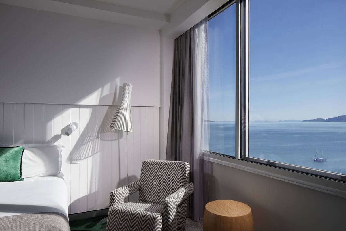A hotel room with a bed, a chair, a lamp, a round wooden table, and a large window with a view of the sea.