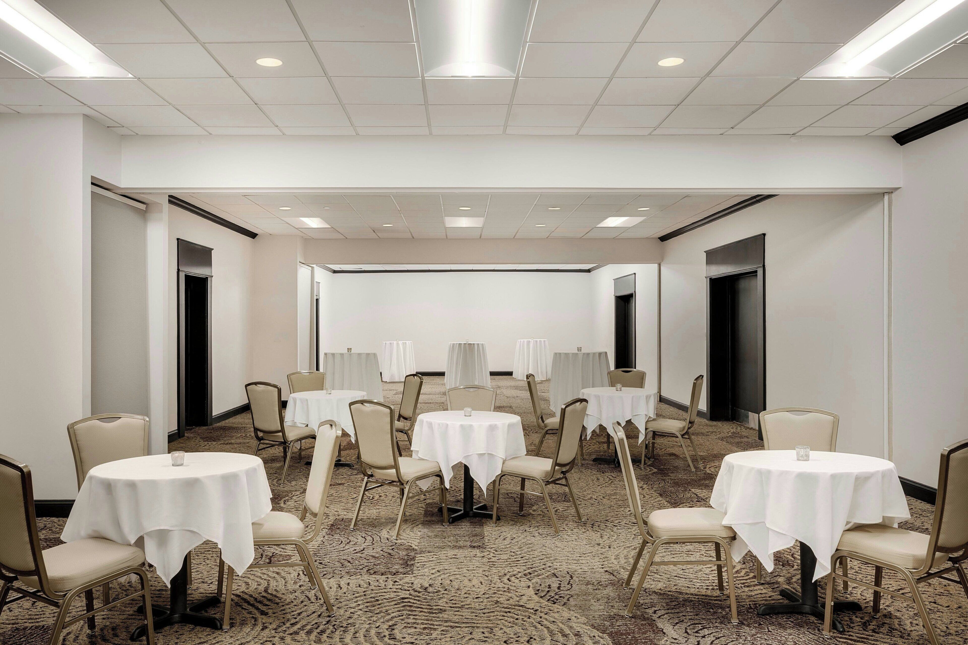 Un salón de conferencias con varias mesas redondas, cada una con mantel blanco y sillas color beige, dispuestas en forma de U alrededor de una pizarra central.