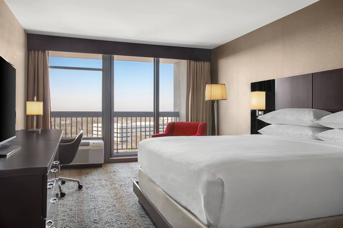 Ein Hotelzimmer mit einem großen Bett, einem Schreibtisch mit Stuhl, einem Fernseher und einem Balkon mit Vorhängen.