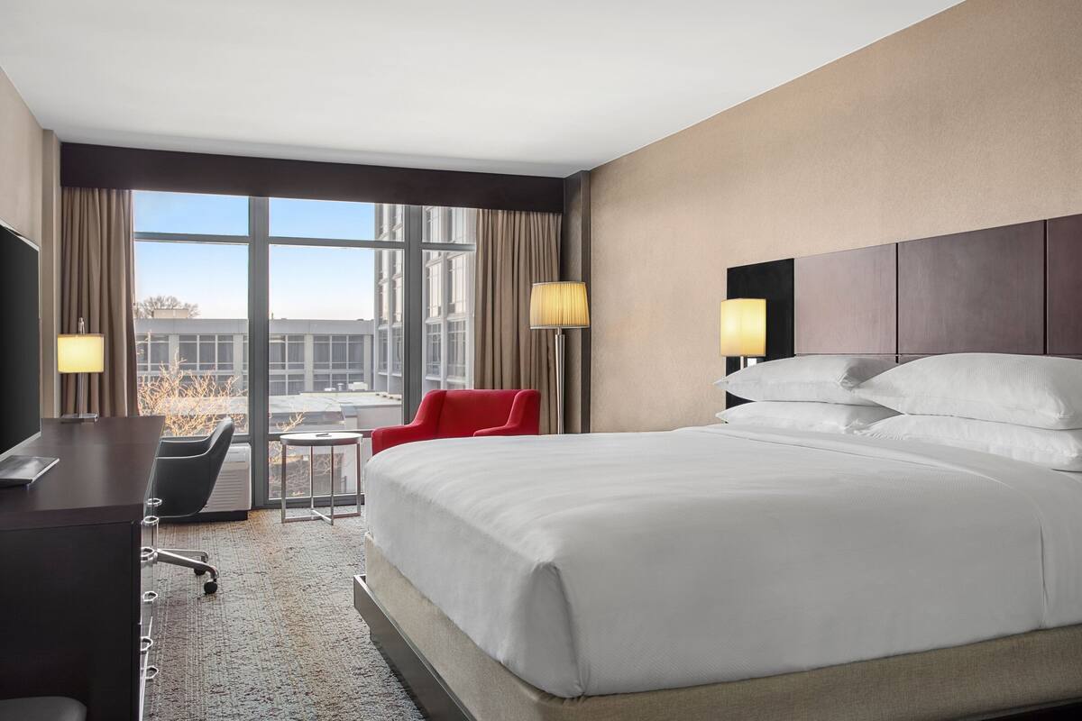 Ein Hotelzimmer mit einem großen Bett, einem Schreibtisch, einem Stuhl und Blick auf die Stadt durch große Fenster.