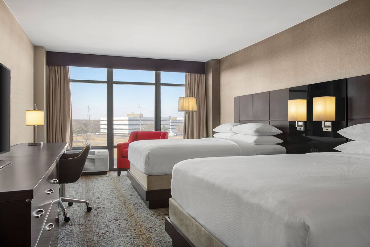 Ein Hotelzimmer mit zwei Betten, einem Schreibtisch, einem Sessel und einem großen Fenster mit Blick auf die Stadt.