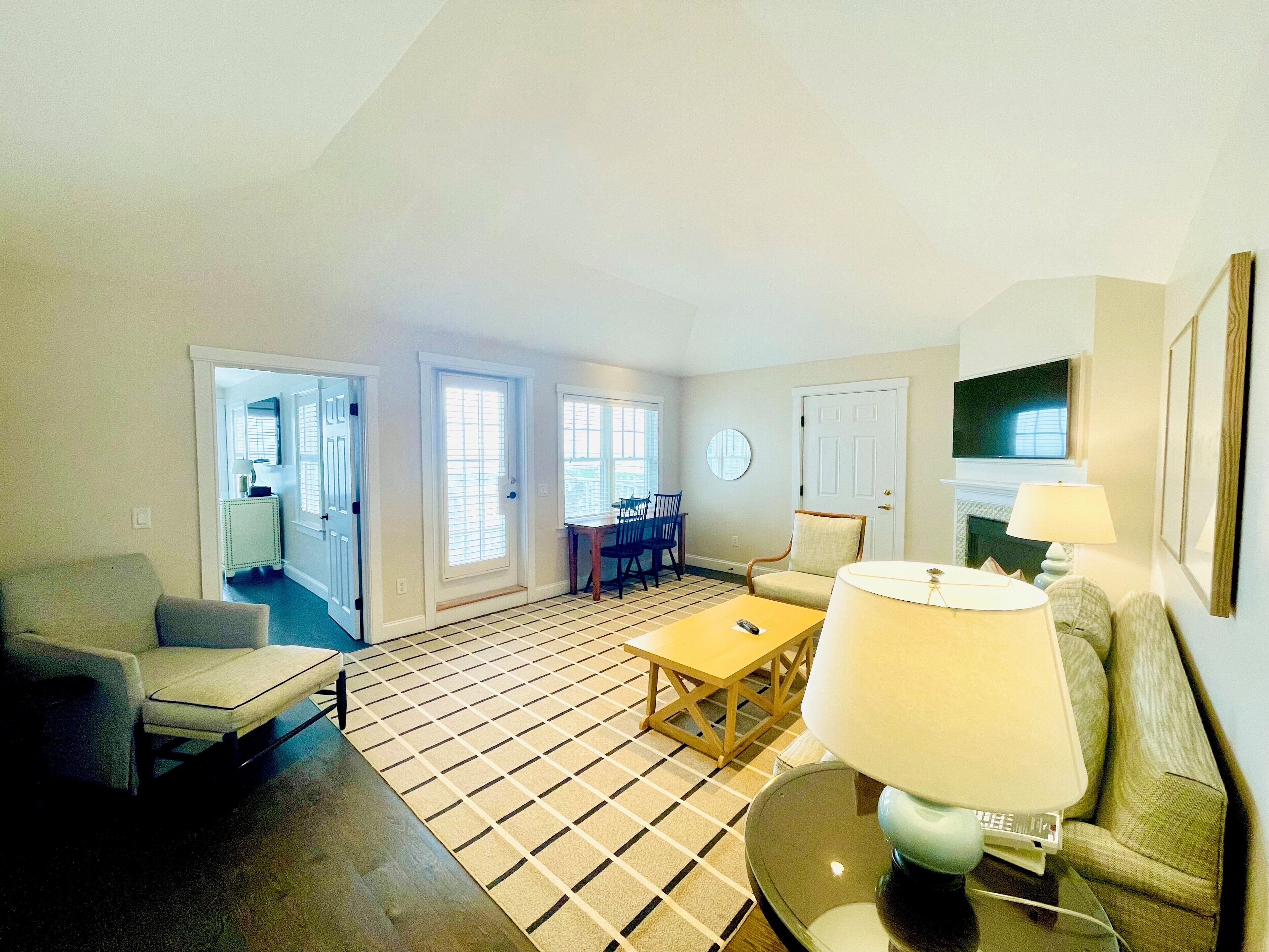 A living room with a sofa, a coffee table, a lamp, and a TV. There is a dining area with a table and chairs. The room has a tiled floor and a door leading to another room.