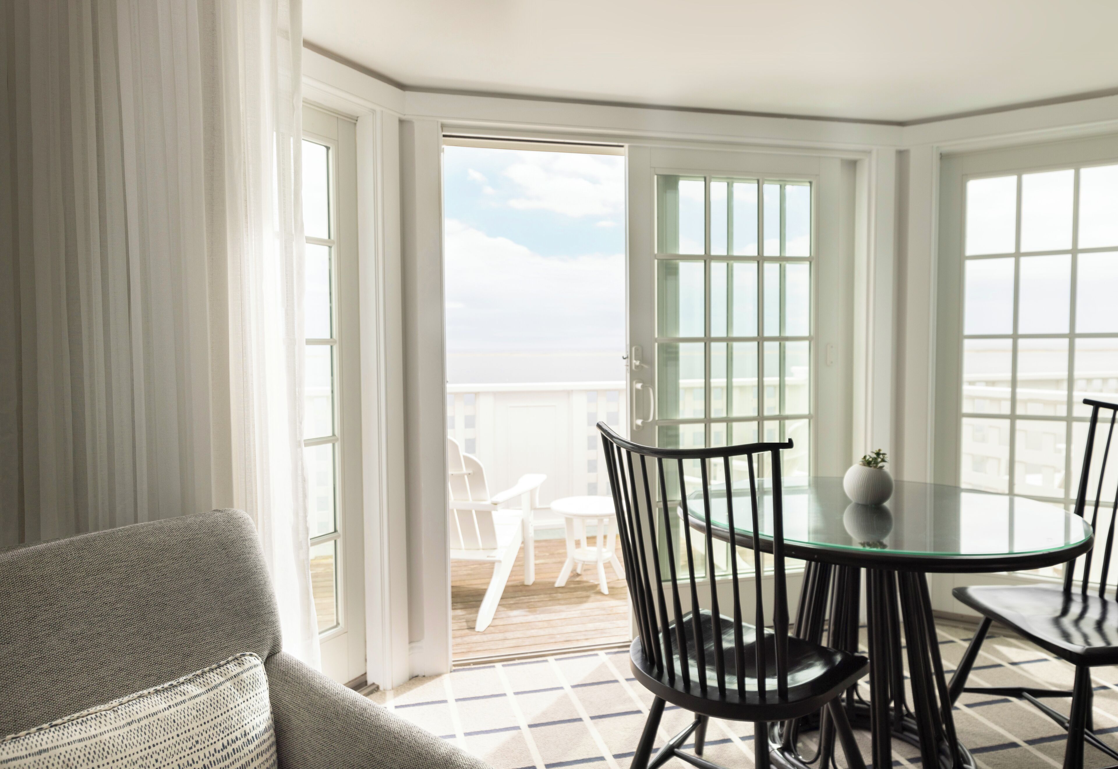 A room with a glass table and chairs, a view of the sea through large windows, and a balcony with outdoor furniture.