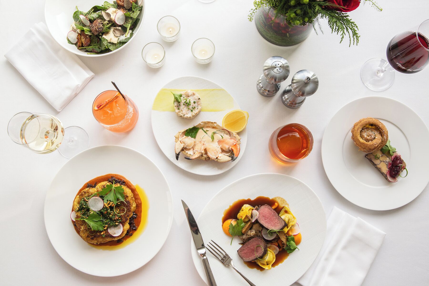 A table set with various dishes, including seafood, a dessert, and a glass of wine, on a white tablecloth.