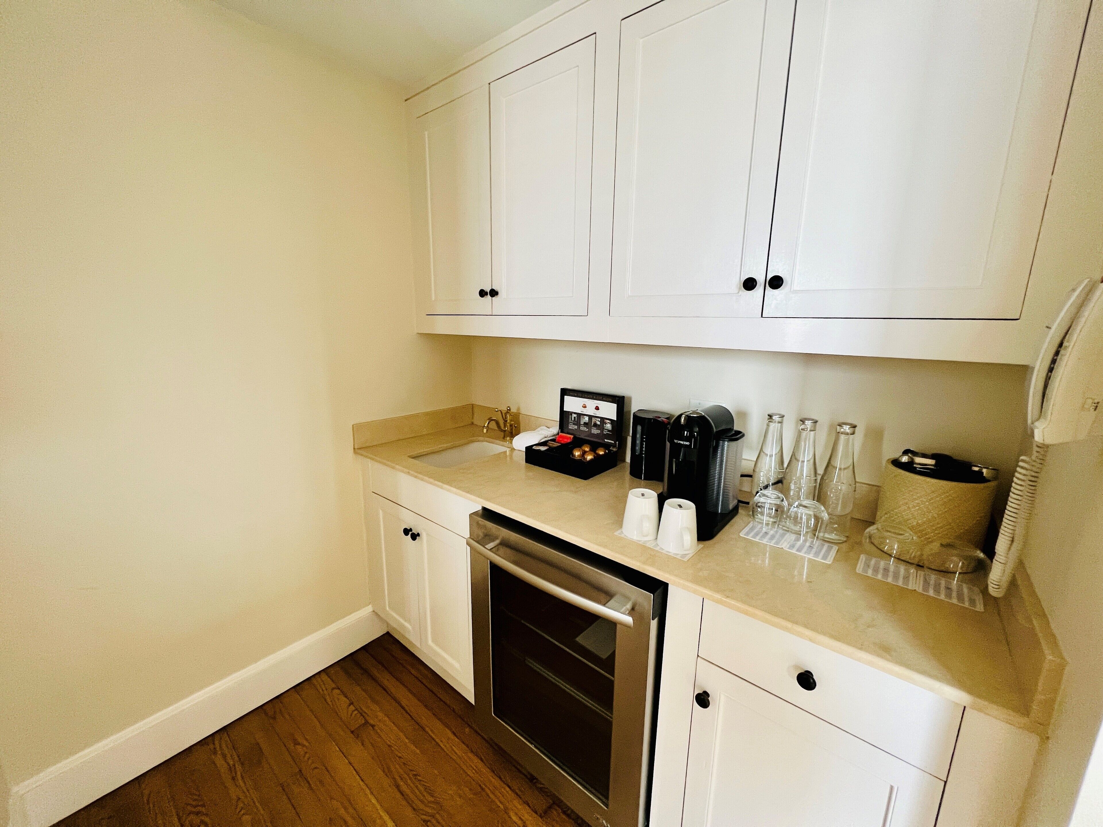 A compact kitchen with white cabinets, a stainless steel dishwasher, a coffee maker, and a wine cooler.