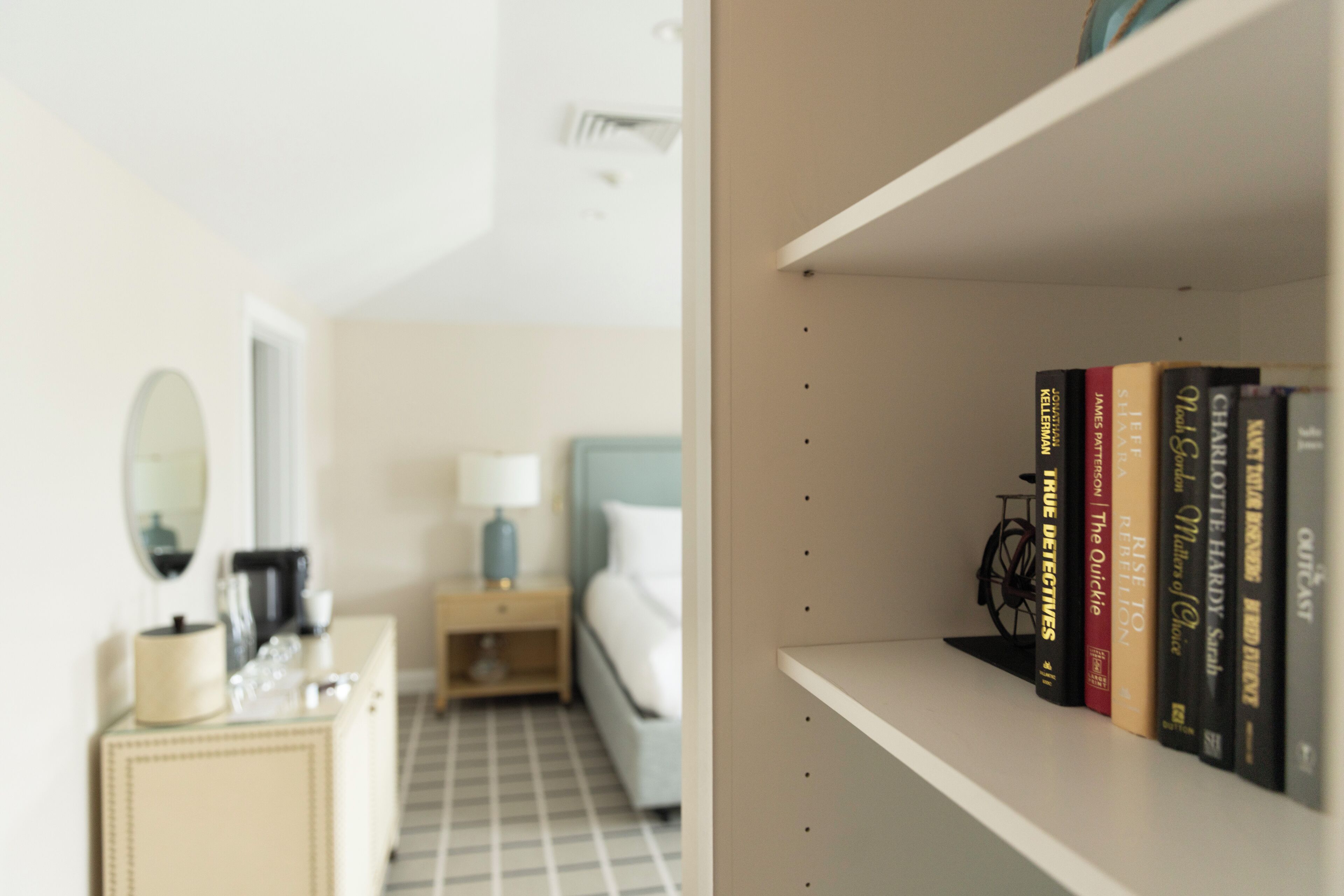 A hotel room with a bed, a nightstand with a lamp, a dresser, and a bookshelf with books.