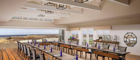 A spacious dining area with long tables and chairs, a wooden floor, and a view of the beach.