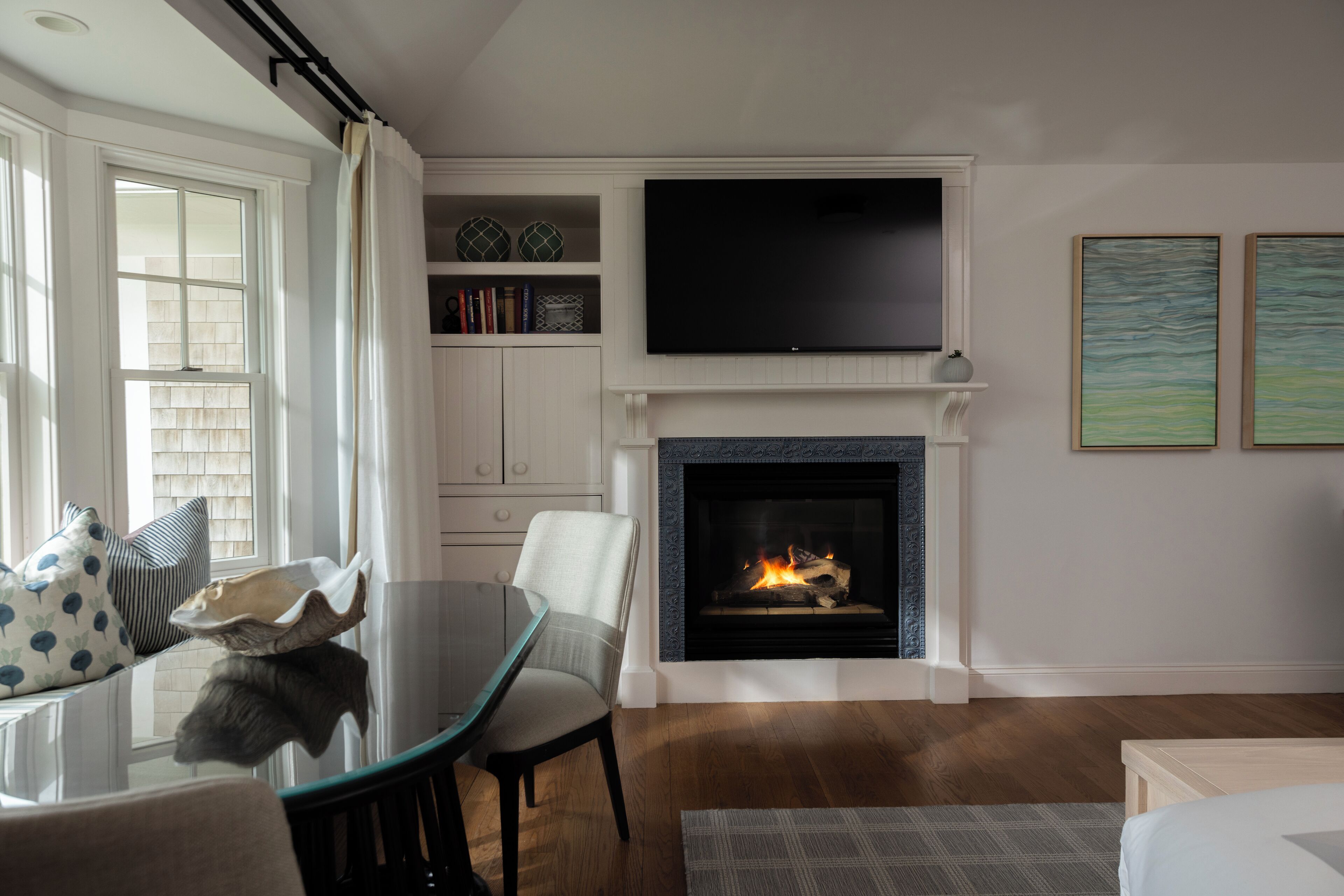 A living room with a fireplace, a flat-screen TV mounted above it, and a seating area with a glass table and chairs.