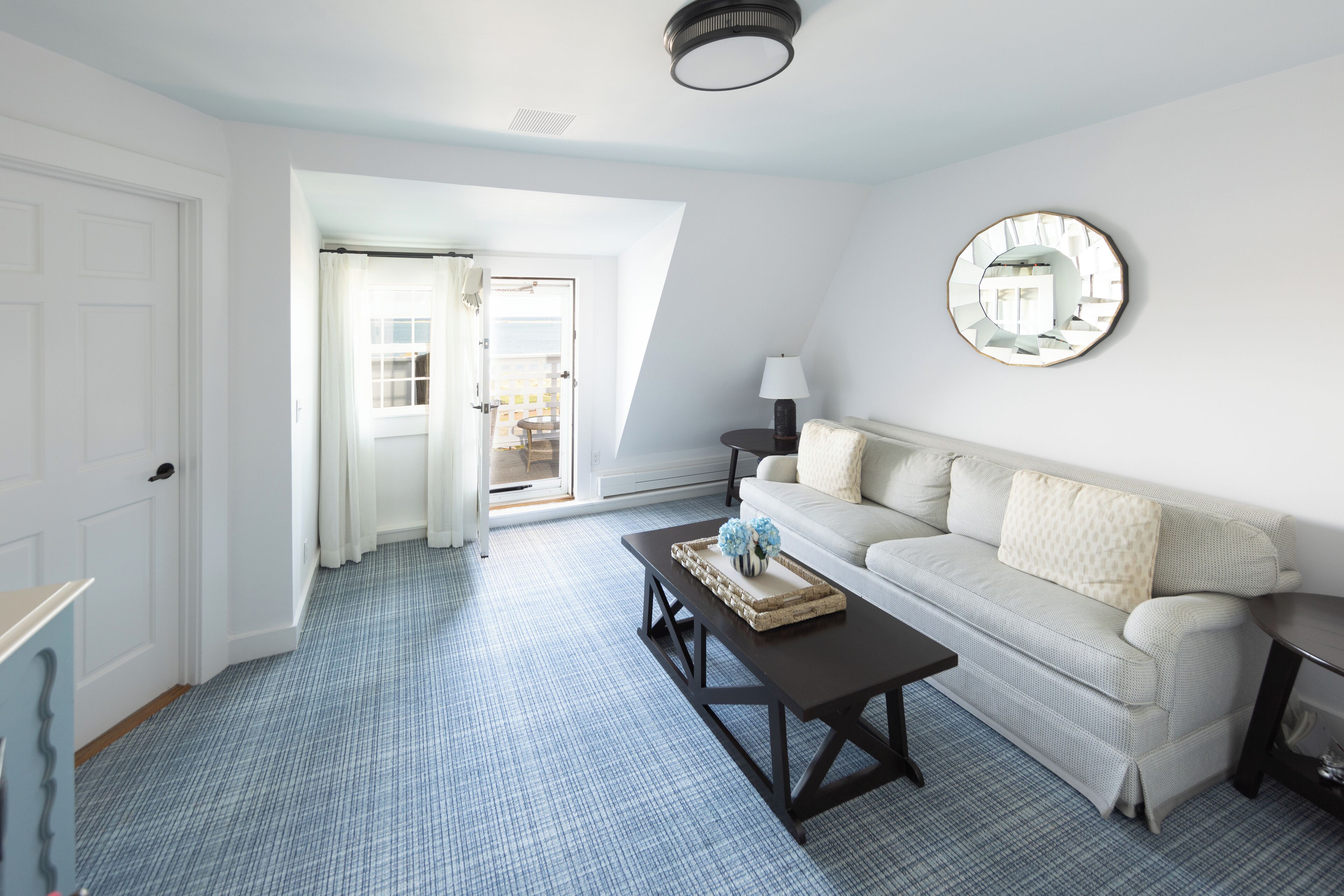 A room with a light-colored sofa, a small round table, a coffee table with a tray, a mirror on the wall, and a door leading to an outdoor area.