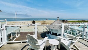 Una terraza con sillas Adirondack blancas y una pequeña mesa redonda, con vista a una playa y al océano.
