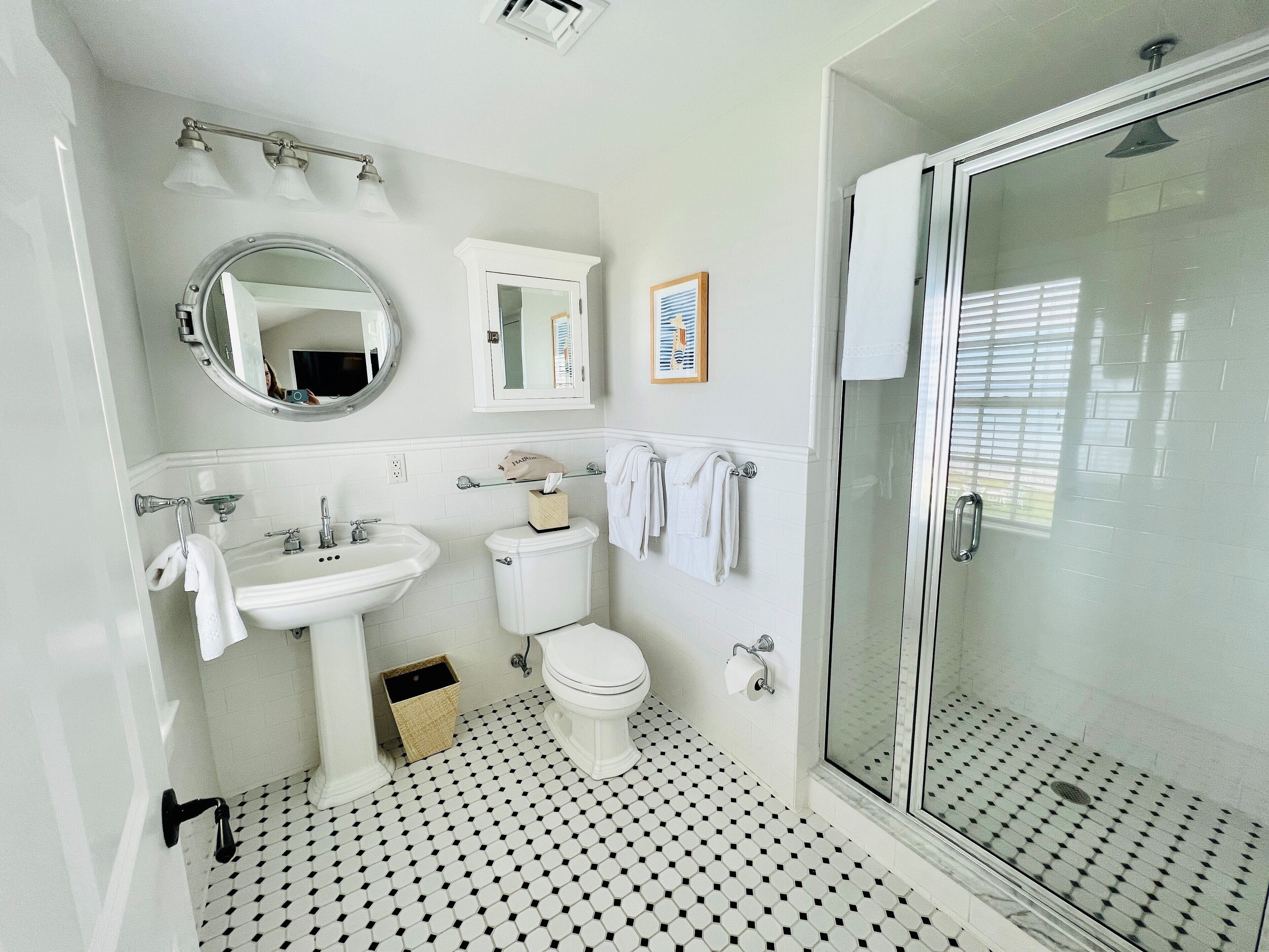 A bathroom with a white sink, toilet, and shower. There is a mirror above the sink and a framed picture on the wall.