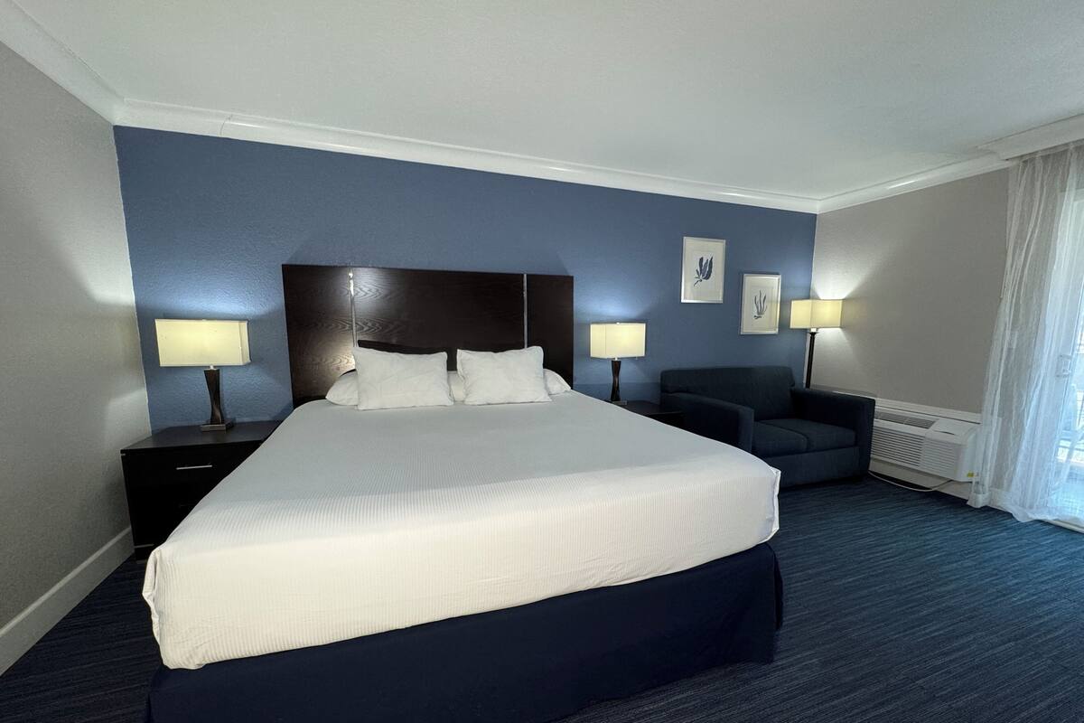 Une chambre d’hôtel avec un grand lit, deux tables de chevet avec des lampes, un fauteuil bleu foncé et un radiateur.
