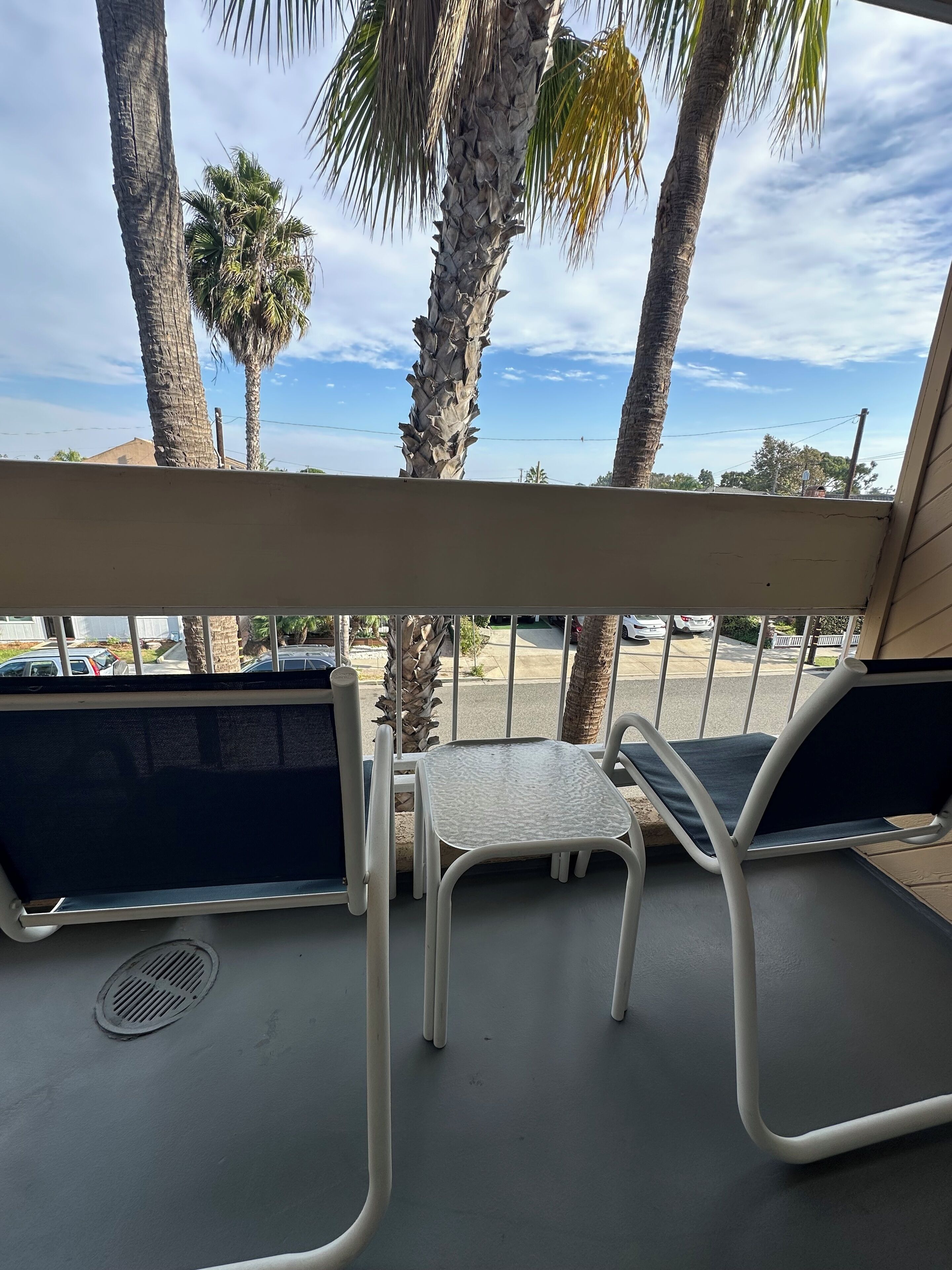 Un balcon avec deux chaises en métal blanc et une petite table, donnant sur un parking et des palmiers.