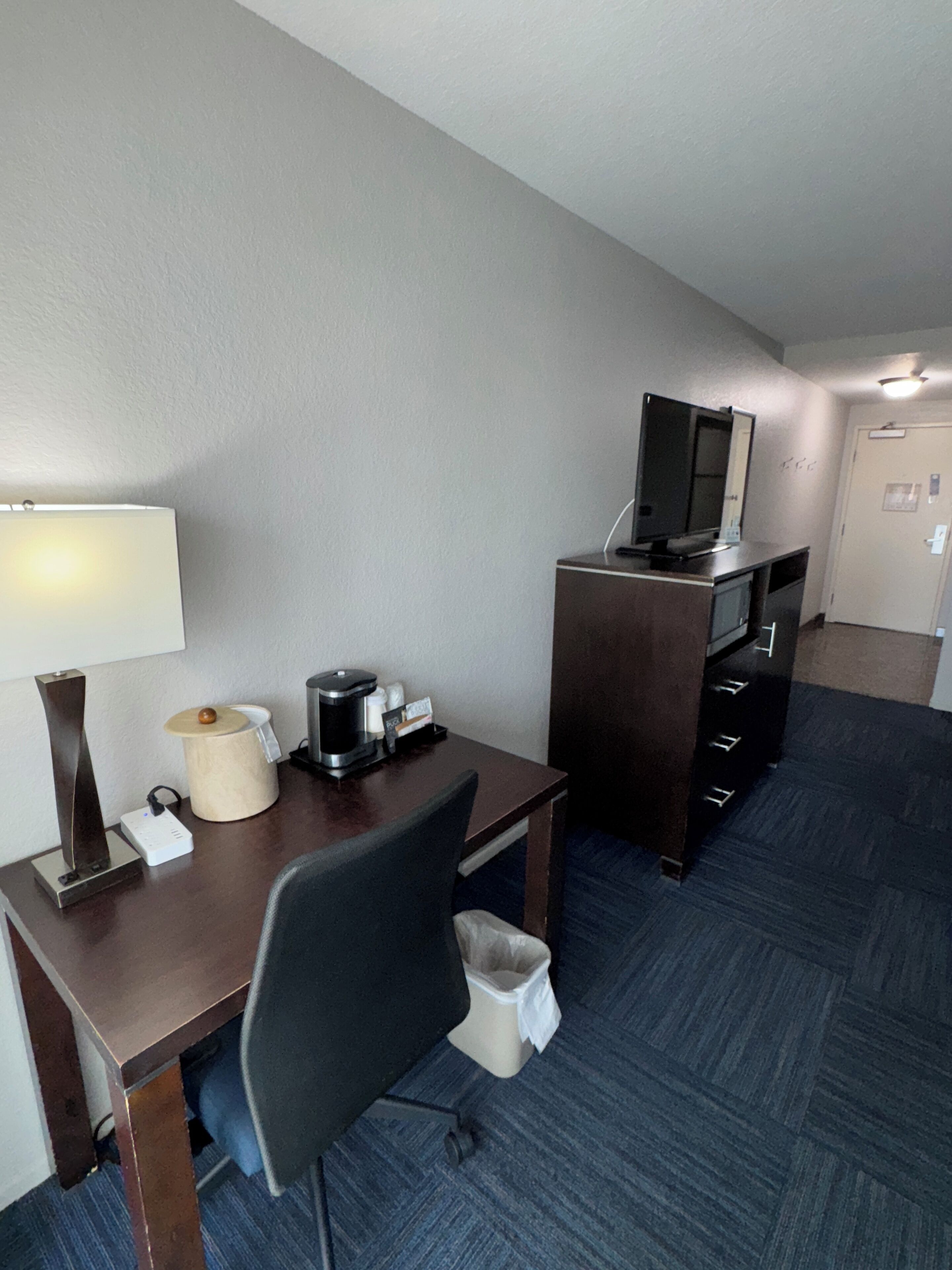 A hotel room with a desk, chair, coffee maker, and a television on a stand.