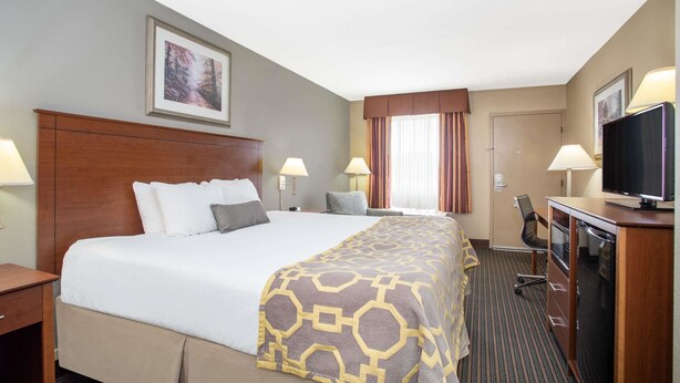 A hotel room with a large bed, two bedside tables, a desk with a chair, a television, and a window with curtains. - Baymont by Wyndham Hays (Hays)