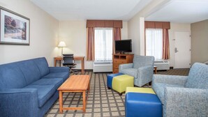 A hotel room with a blue sofa, a wooden coffee table, a television on a stand, and a small desk with a lamp. - Baymont by Wyndham Hays (Hays)