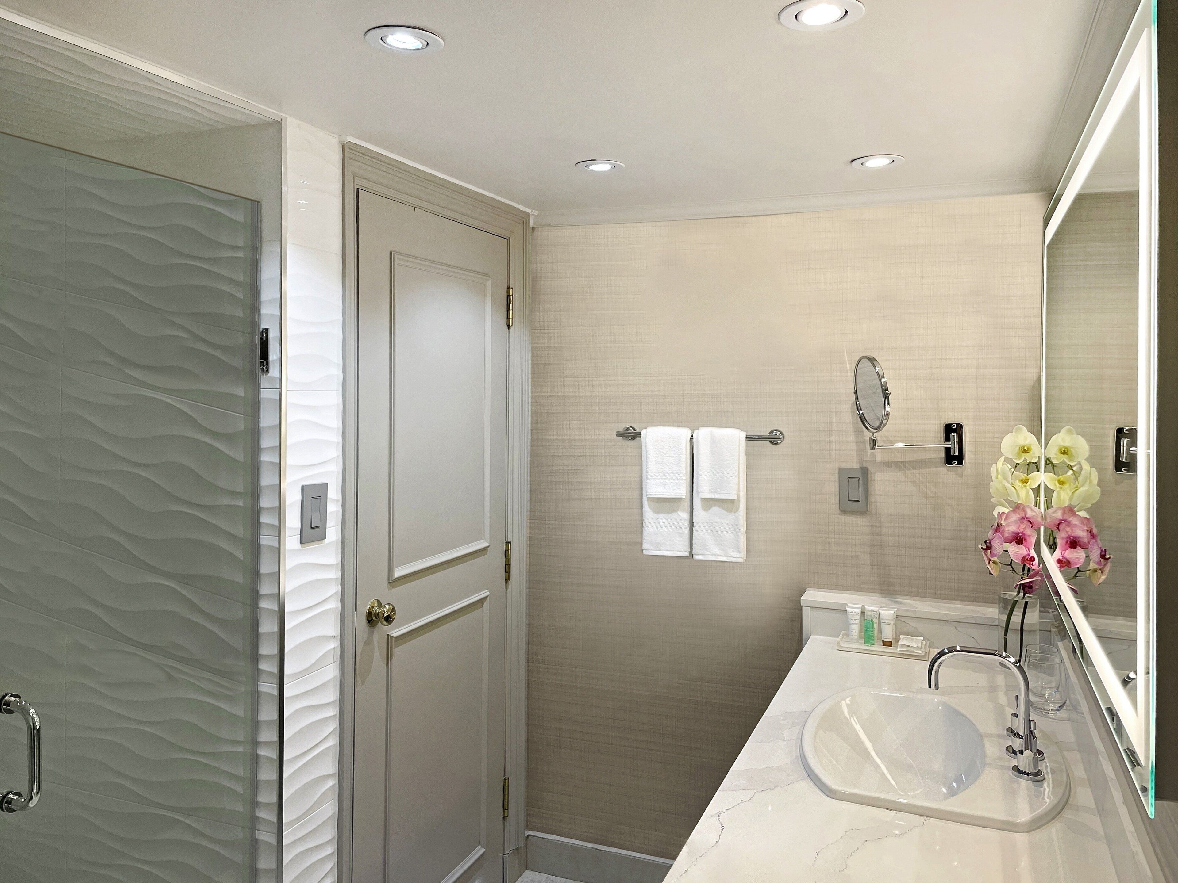 Un baño moderno con cabina de ducha de vidrio, lavabo de mármol, toallero con dos toallas blancas, espejo y un florero con flores.