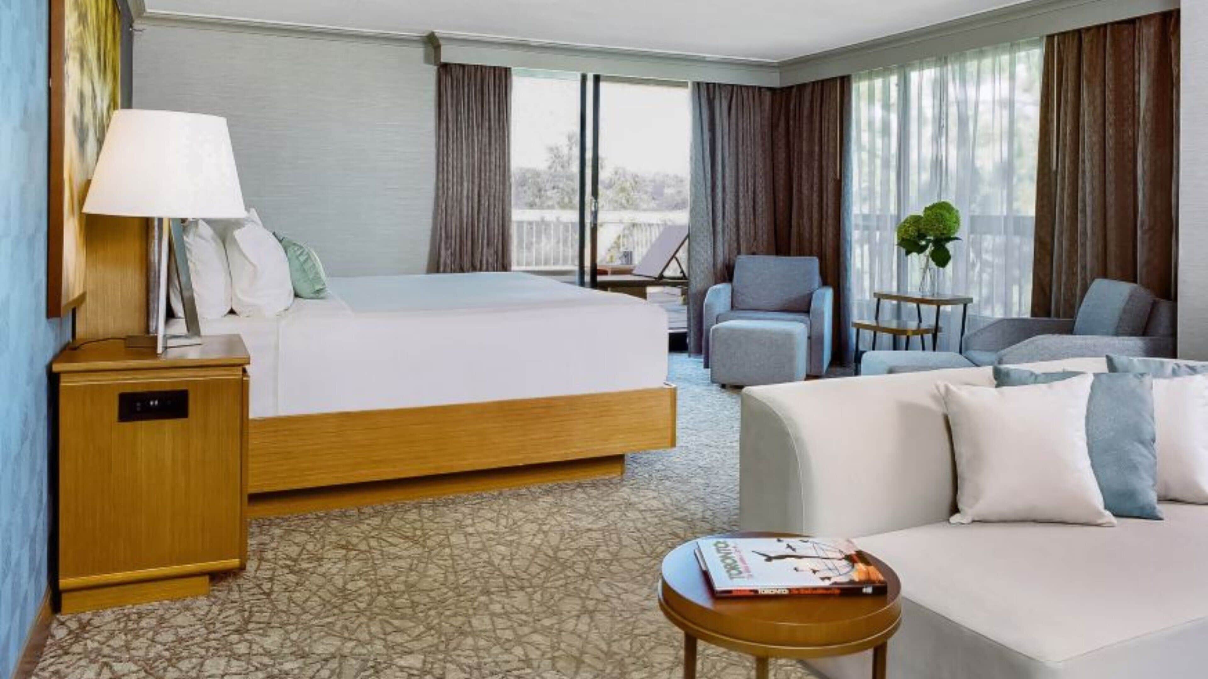 A hotel room with a bed, two armchairs, a sofa, a small table with a book, a lamp, and a window with curtains.