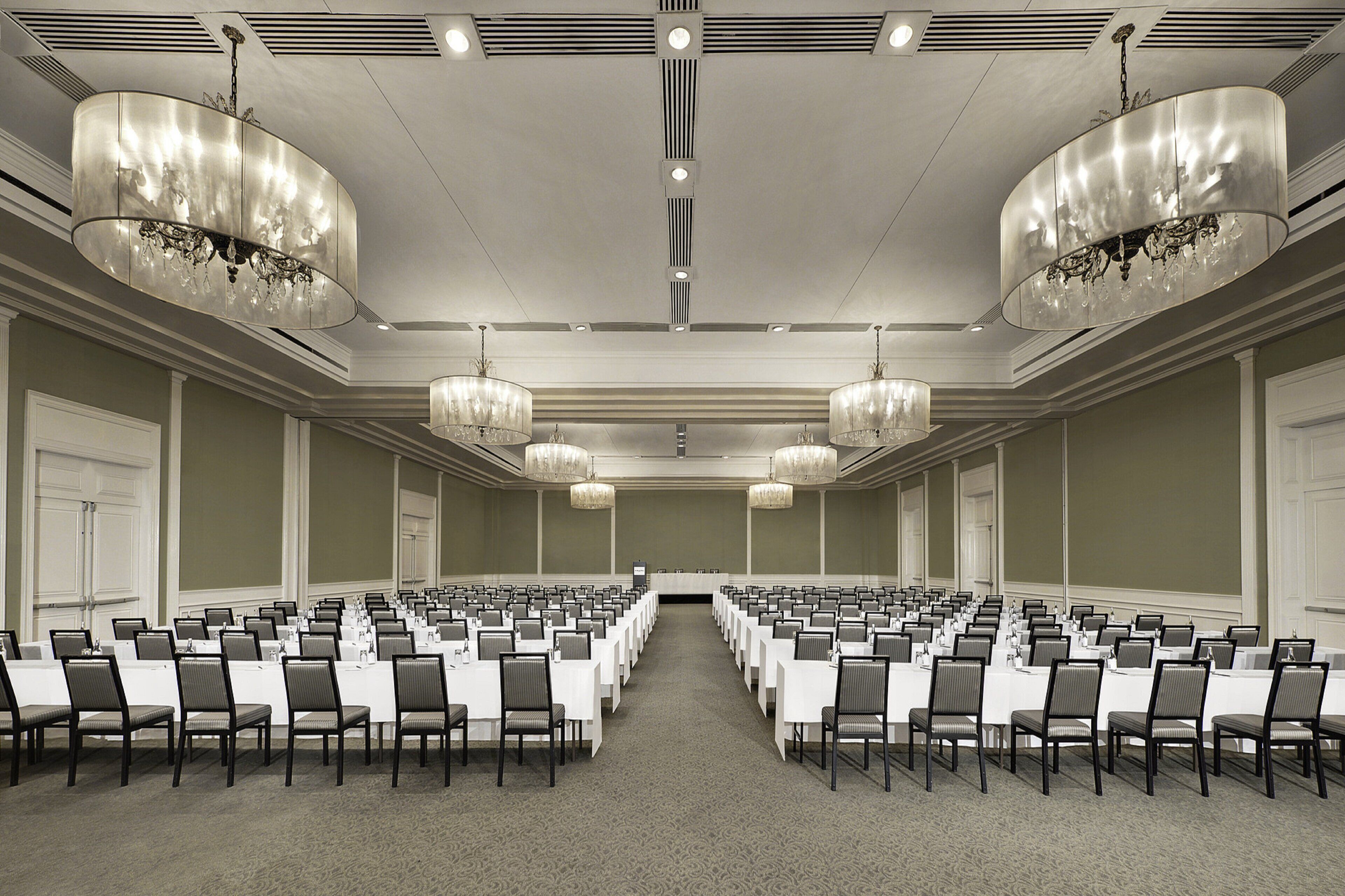 A conference room with rows of tables and chairs, large chandeliers, and a carpeted floor.