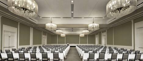 A conference room with rows of tables and chairs, large chandeliers, and a carpeted floor.