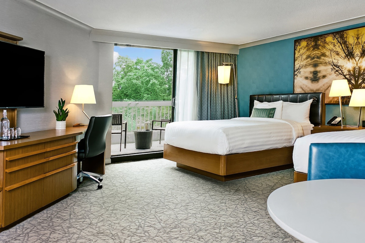 A hotel room with a bed, a desk, a chair, a television, and a window with a view of trees.