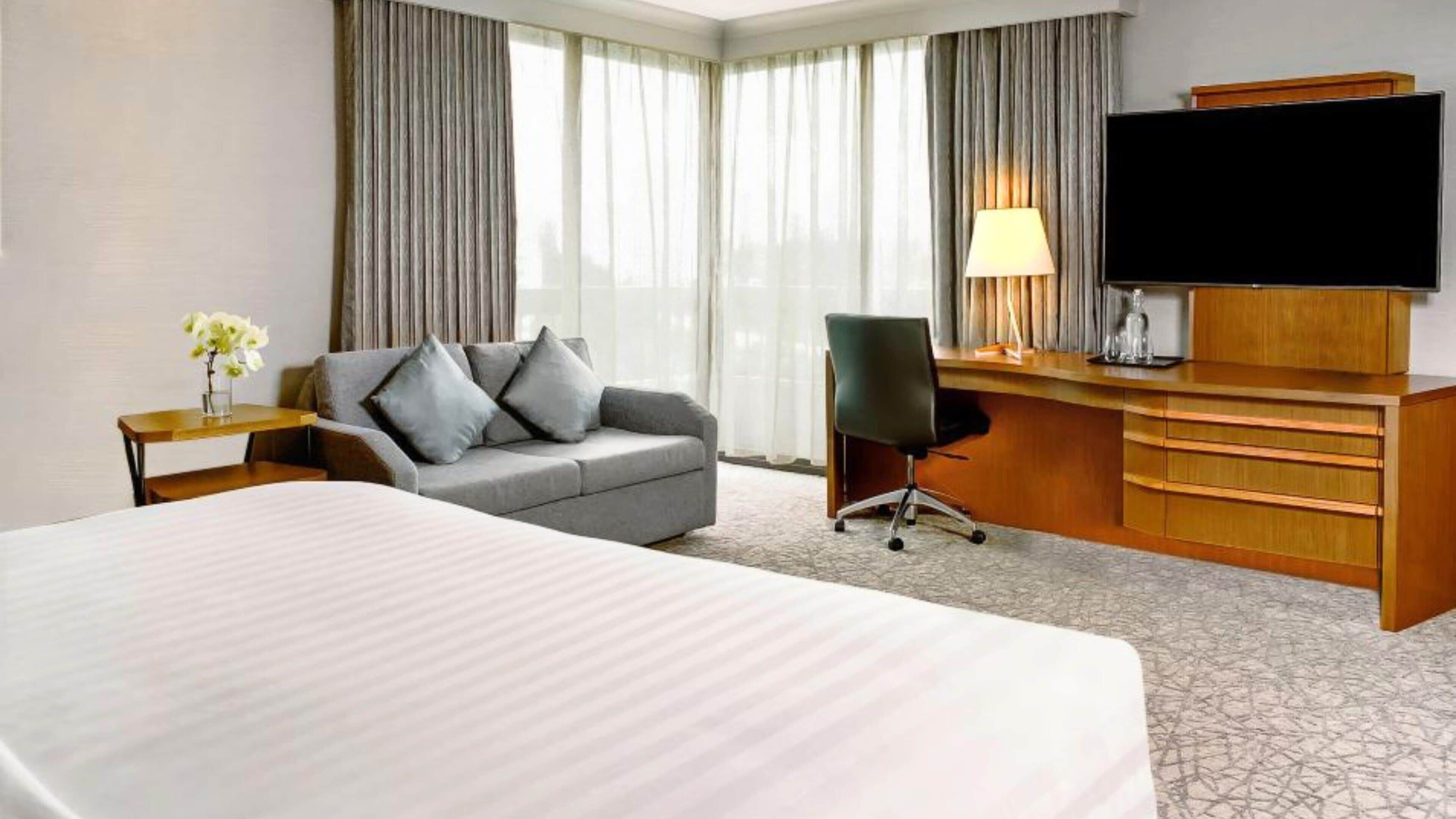 Et hotelværelse med en stor seng, en grå sofa, et træskrivebord med et fjernsyn og en lampe.