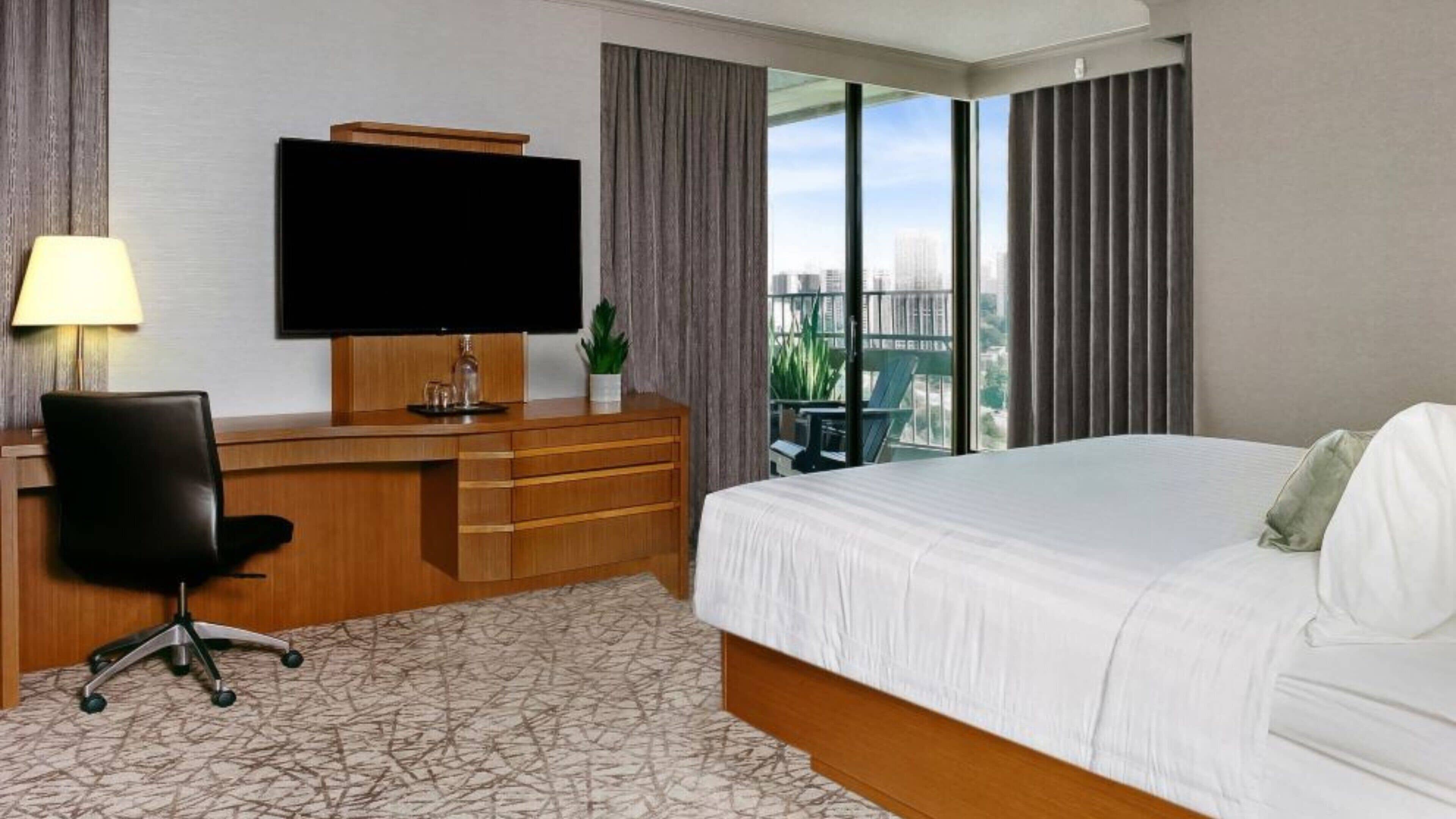 A hotel room with a bed, a desk with a chair, a TV, a plant, and a view of a cityscape.