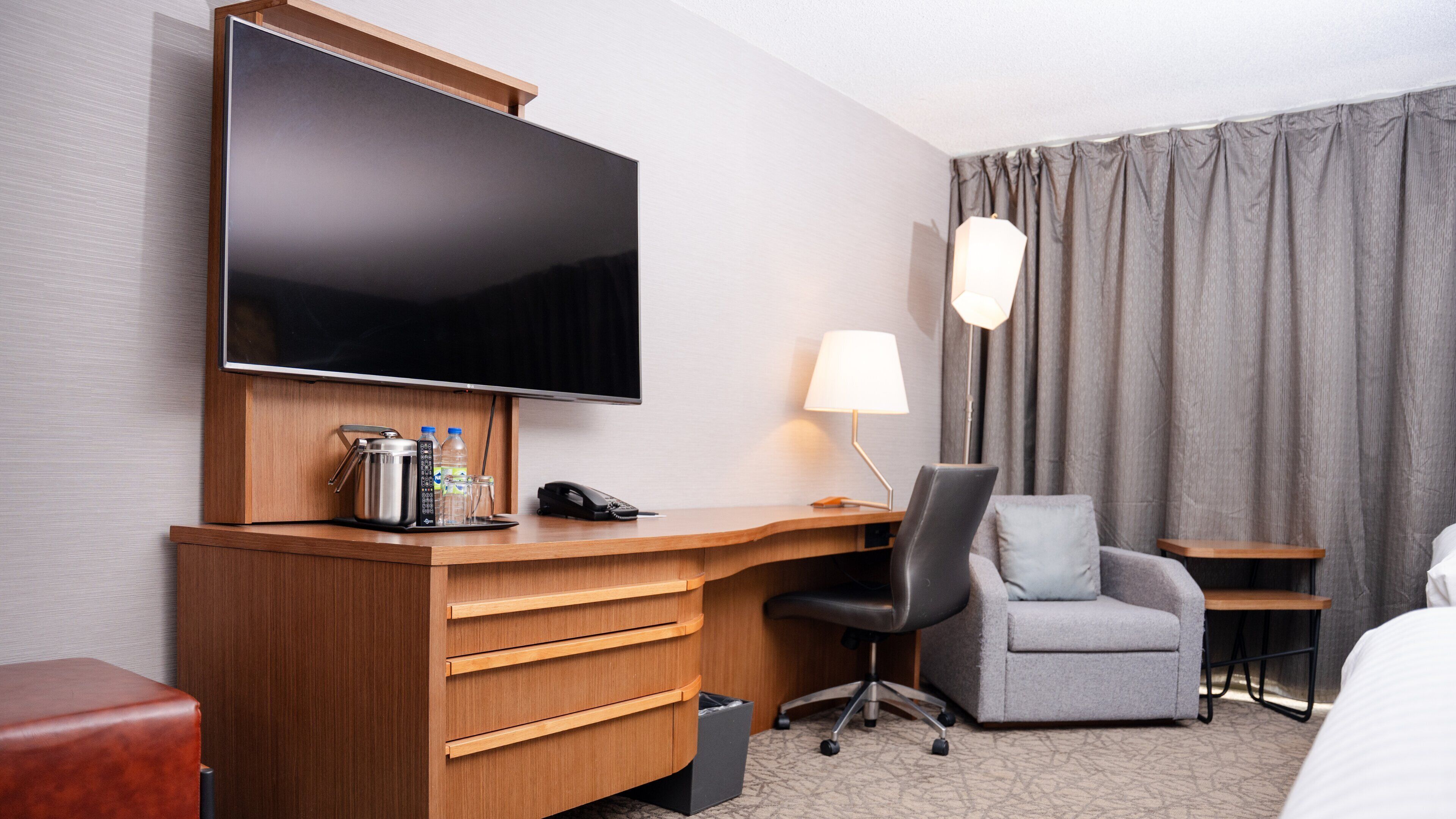 A hotel room with a wooden desk, a flat-screen TV, a grey armchair, a small bedside table, and a lamp.