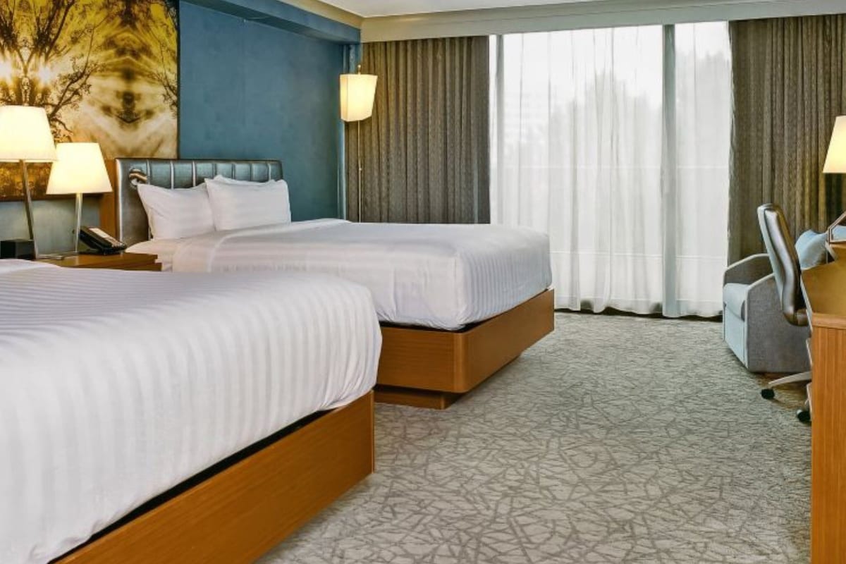 A hotel room with two beds, a desk, a chair, a TV, and a large window with curtains.