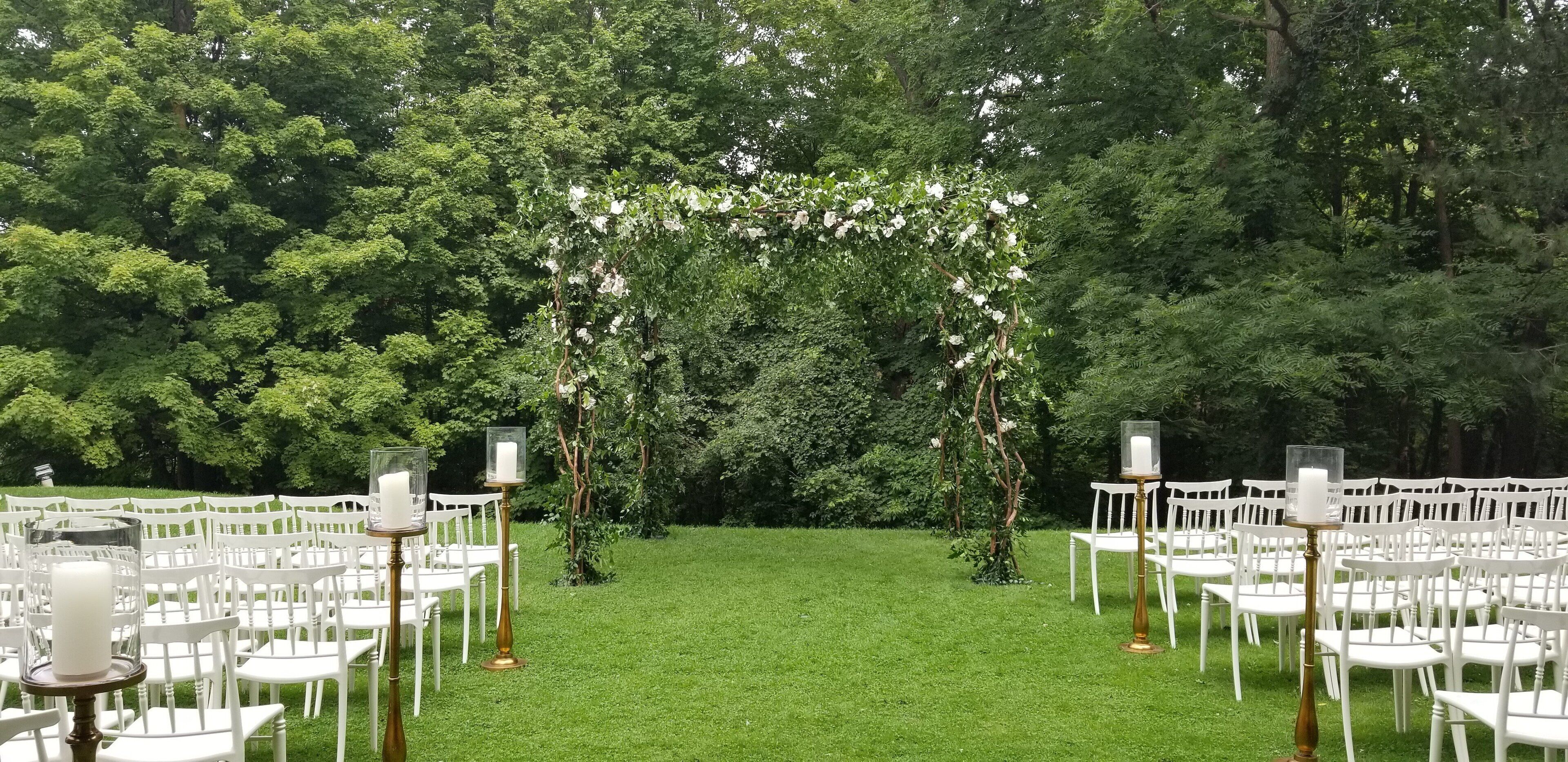 Un salón de bodas al aire libre con sillas blancas dispuestas en filas, un arco floral central y un exuberante fondo verde.