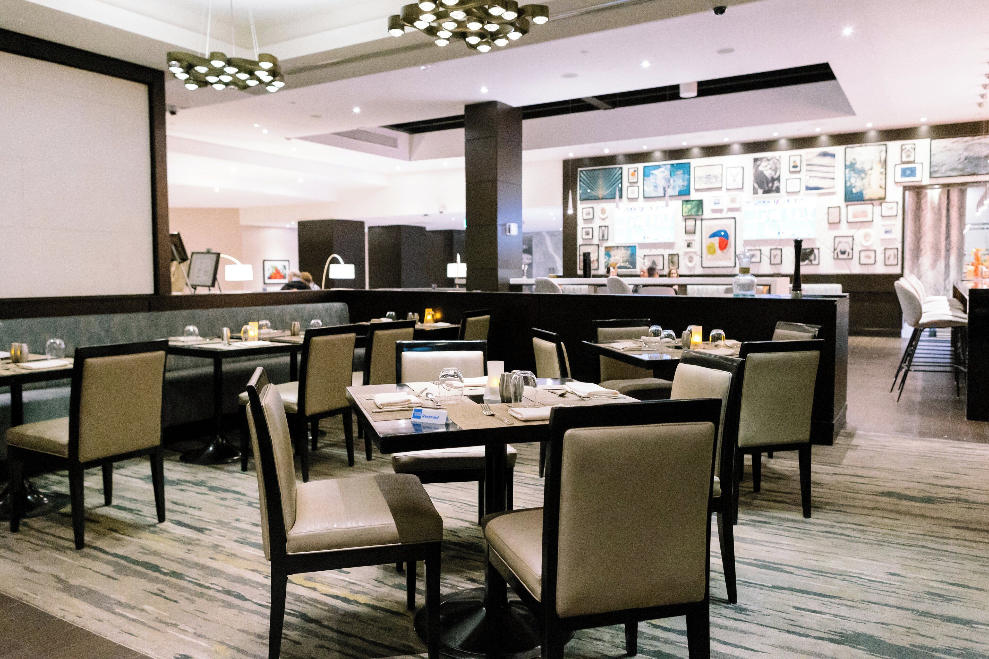 A modern hotel lobby with multiple dining tables and chairs, a reception desk, and a wall adorned with framed pictures.