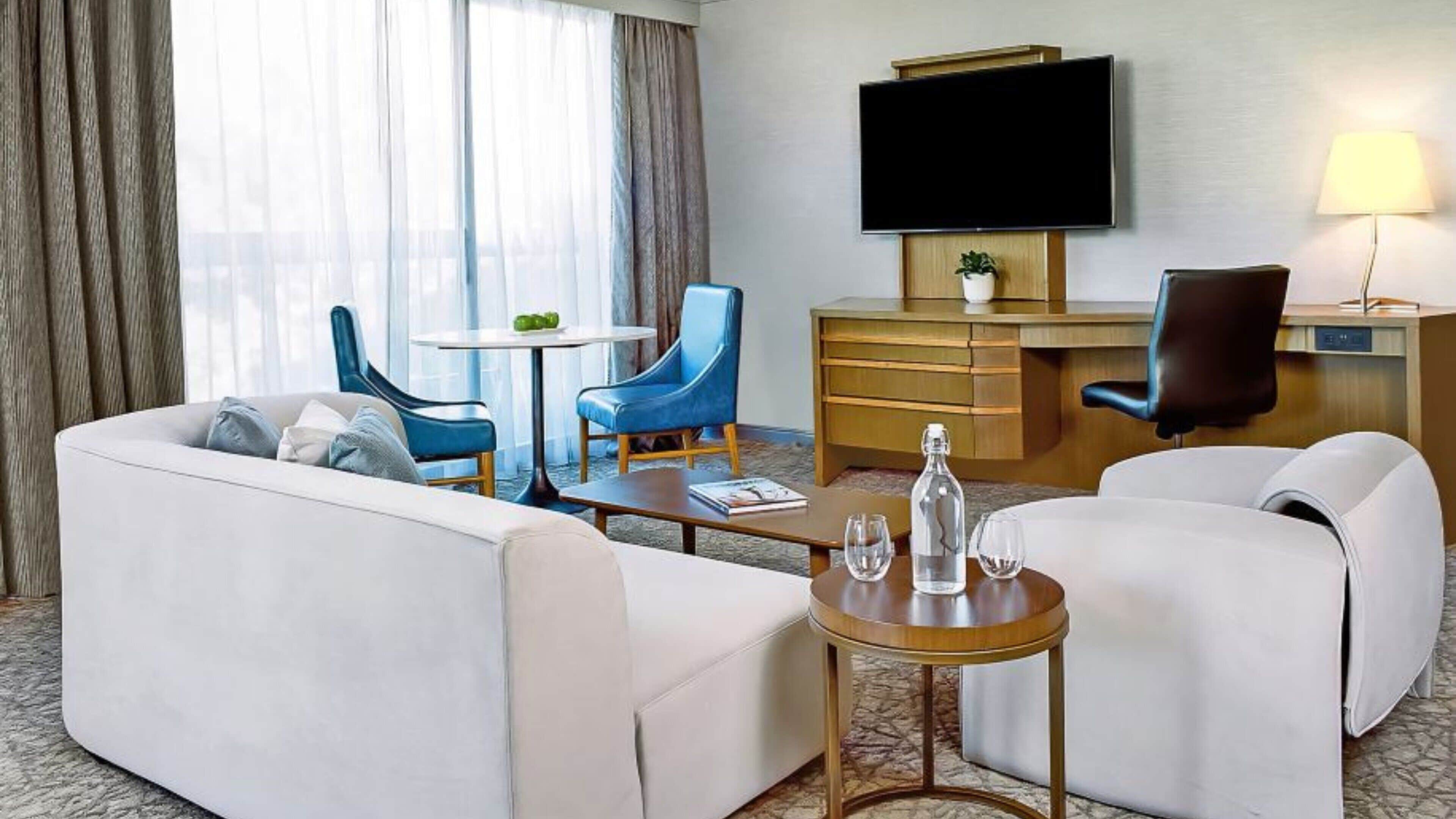 Quarto de hotel com sofá, duas poltronas, uma mesa pequena, televisão, escrivaninha e abajur.