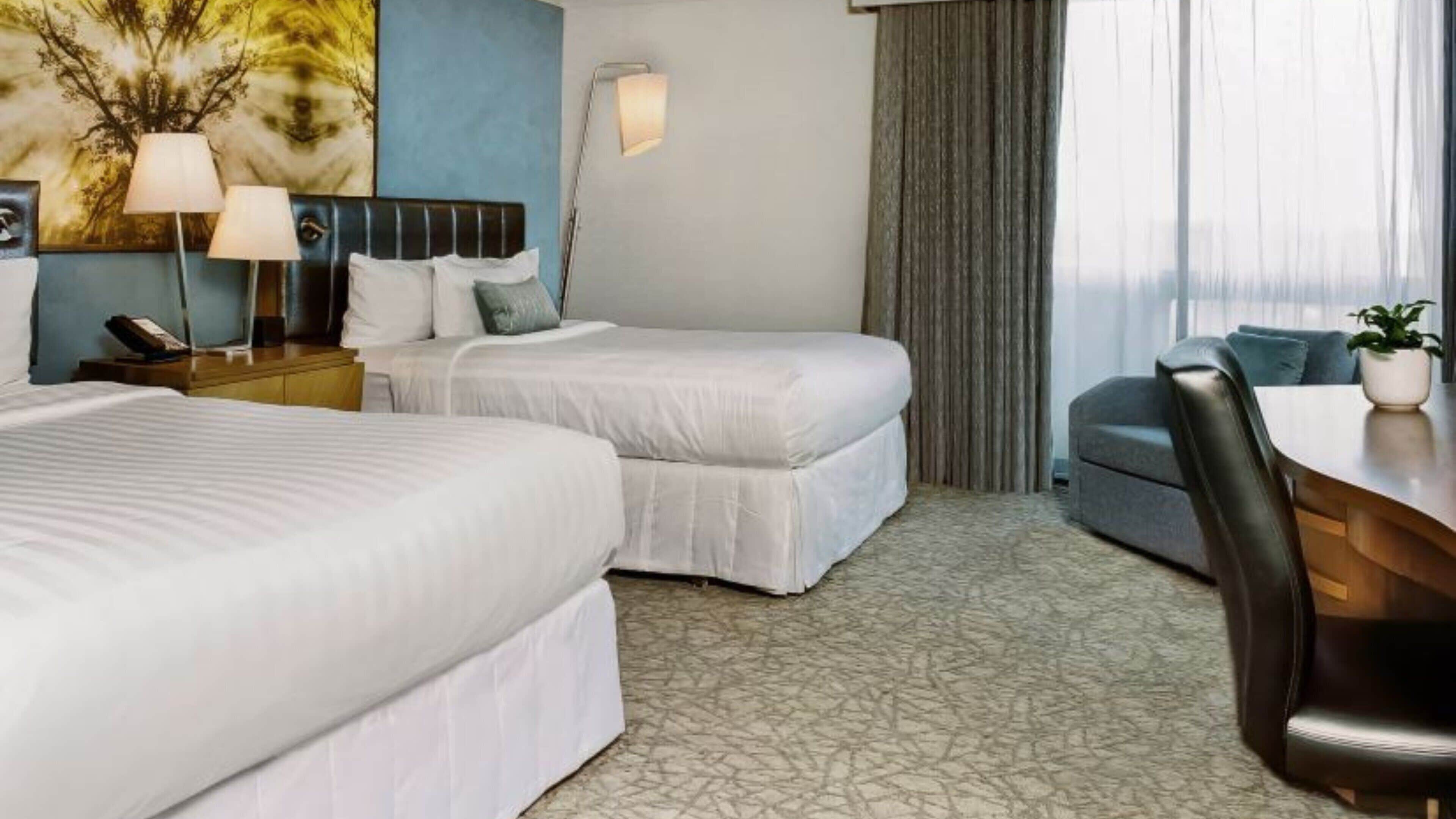 Quarto de hotel com duas camas, uma mesa, uma cadeira, uma luminária e uma janela grande com cortinas.